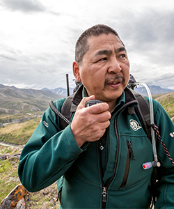 Un employé de Parcs Canada parle sur une radio bidirectionnelle, des champs et des montagnes en arrière-plan.