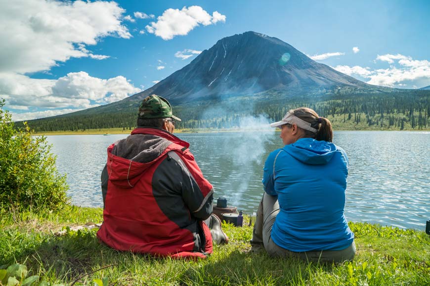 Un homme et une femme sont assis devant un lac pendant que la vapeur monte devant eux à partir d'un pot avec une grande montagne directement en face.
