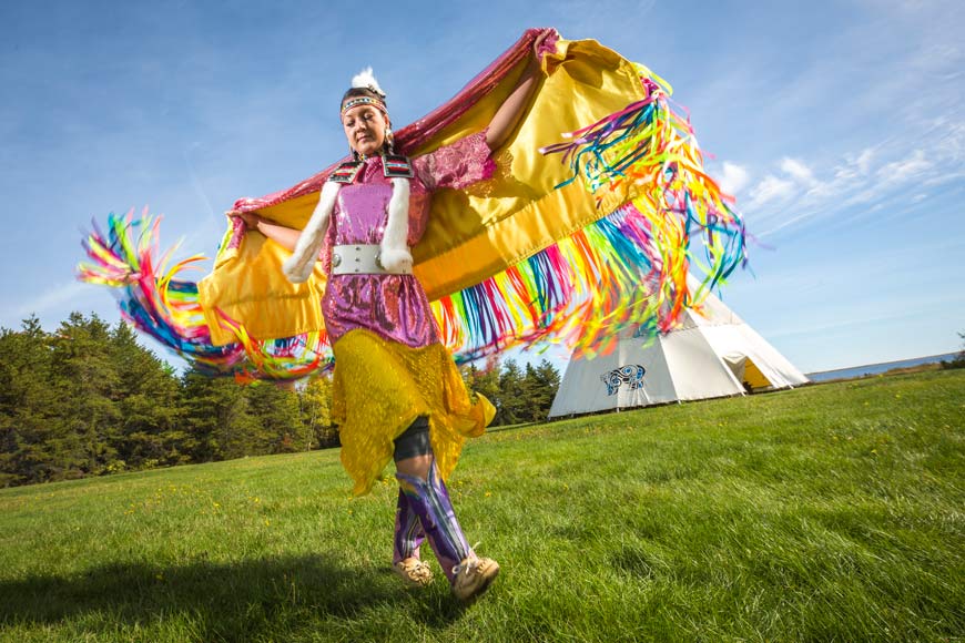 Femme autochtone portant une châle fantaisie, dansant dans l'herbe verte devant un grand Wigwam.