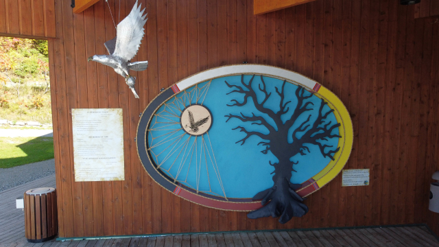 Sur l'extérieur d'un bâtiment, une œuvre d'art circulaire avec du blanc, du jaune, du rouge et du noir sur la bordure du cercle. Au centre, un fond bleu, un arbre noir et l'image d'un oiseau relié par des cordes. Un oiseau argenté est suspendu près de l'œuvre d'art.