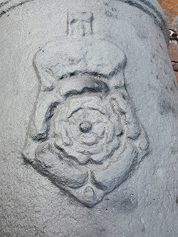 Gros plan d’une plaque en relief sur un canon. Il s’agit d’un monogramme surmonté d’une couronne, en haut, et d’un pentagone, en bas, avec une rose au milieu.