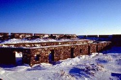 Un mur de pierre à l’arrière-plan de l’image sert de mur extérieur au fort Prince-de-Galles, tandis que deux murs de pierre plus courts, plus proches de l’observateur, sont des murs intérieurs avec des portes et des fenêtres. Le sol est recouvert de neige.