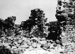 Une image en noir et blanc montre des pierres au premier plan. À droite, il y a un mur de briques dont le bord est dentelé, et au centre, une autre section du mur de briques, plus loin de l’observateur.