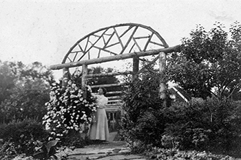 Une femme debout un chemin pavé de sous une arche en bois elle touche les fleurs