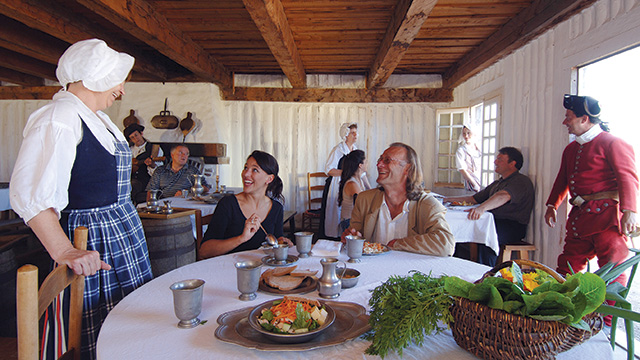 Deux personnes sont assises à une table recouverte de lin et s'entretiennent avec une femme habillée comme au XVIIIe siècle. Sur la table se trouvent du pain frais, une salade et un panier d'herbes fraîches.