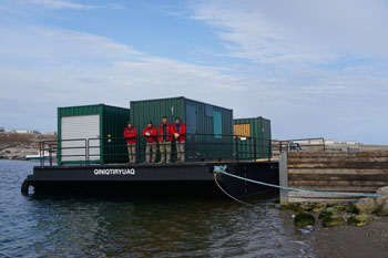 Quatre personnes vêtues en rouge debout sur une barge devant des unités de stockage en métal vert.