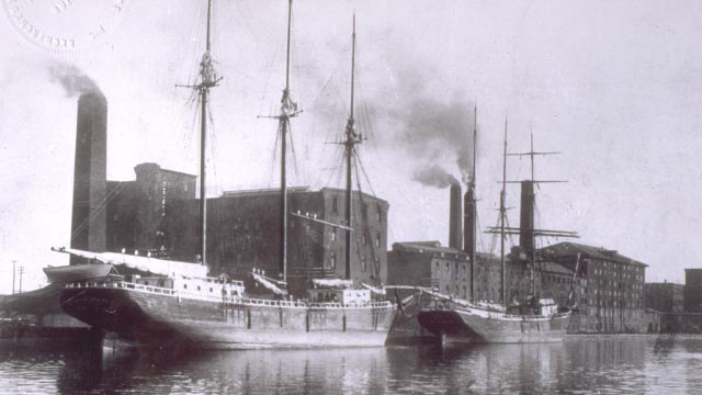 Image ancienne en noir et blanc d'un bateau dans un canal devant un bâtiment industriel