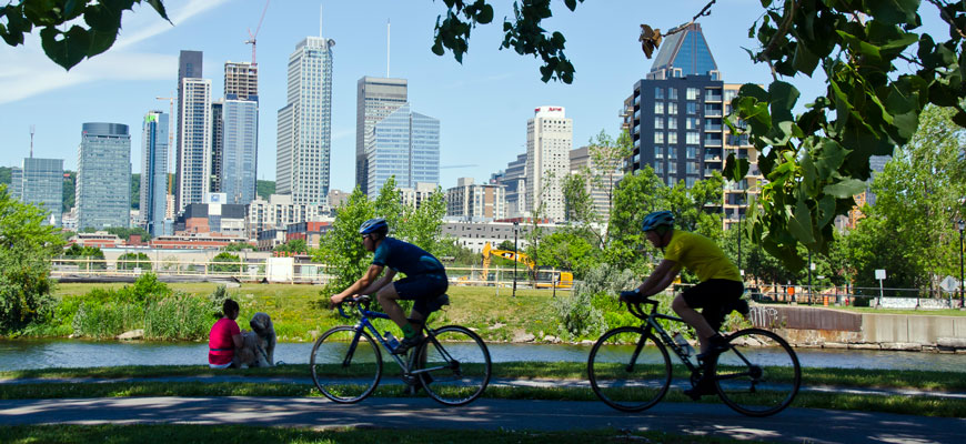 Des visiteurs font du vélo le long du canal de Lachine avec le centre-ville de Montréal en arrière-plan.