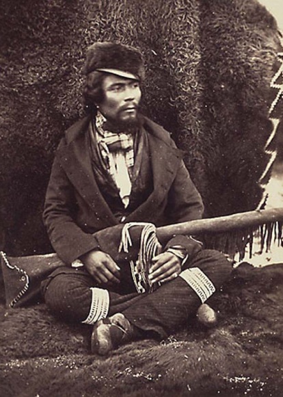 Photo ancienne d'un homme assis, les jambes croisées, contre un tas de peaux de bison, tenant son fusil.