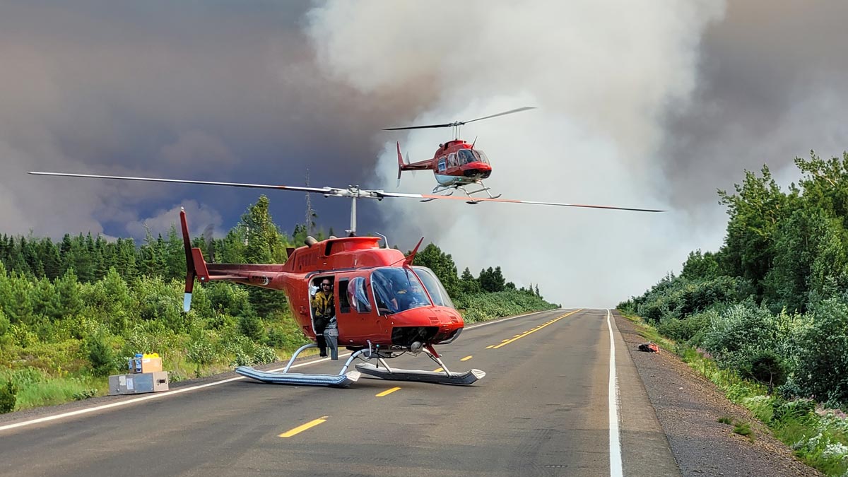 Un hélicoptère se trouve sur une autoroute fermée. En arrière plan, on aperçoit une épaisse fumée de feu de forêt et un deuxième hélicoptère qui décolle dans les airs.