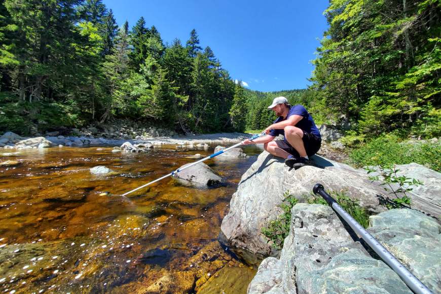 Un chercheur est assis sur un rocher au bord d’une rivière alors qu’il utilise une longue perche dans l’eau pour prélever un échantillon d’eau.