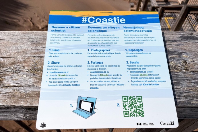 Vue rapprochée d'un panneau fixé à une promenade en bois sur une plage de sable, donnant des instructions pour réaliser une photo pour l'initiative Coastie.