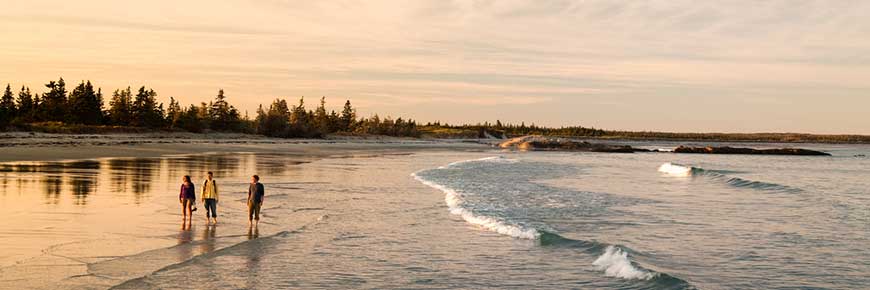 A panoramic view of Kejimkujik Seaside at sunset, with three people to the left in the distance walking along the shoreline. 