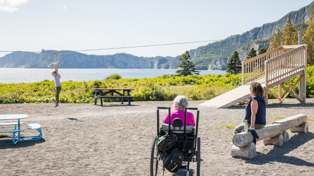 Une enfant joue dans un terrain de jeu accompagnée par une dame en fauteuil roulant et une autre assise sur un banc. 