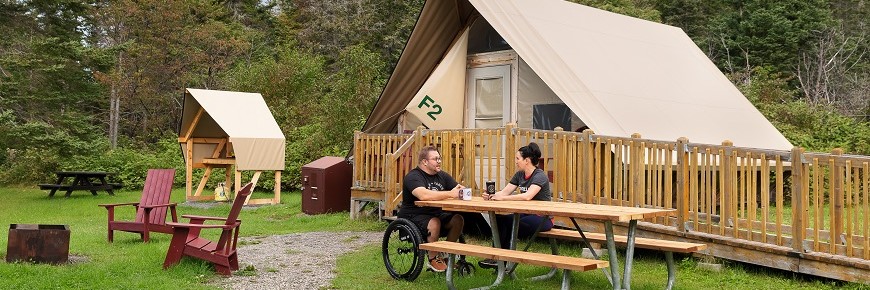 Un homme en fauteuil roulant et une femme sont installés à la table de pique-nique adaptée pour les personnes à mobilité réduite, devant la tente oTENTik F2 du camping Petit-Gaspé. Une rampe permet d'accéder dans la tente. 