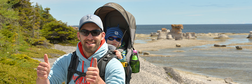 Photographie d'un père avec son enfant dans un porte-bébé avec des monolithes et la mer en arrière-plan
