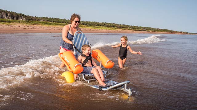 Un enfant excité dans un fauteuil roulant de plage flottant est guidé dans l'eau par un adulte, avec un autre enfant excité à proximité.