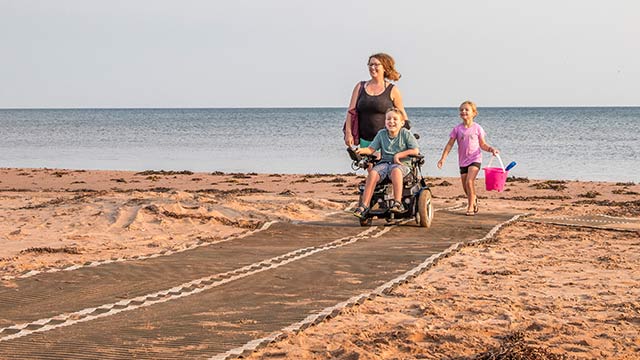 Un enfant en fauteuil roulant, un autre tenant des jouets, et une adulte, reviennent de la plage en utilisant un tapis de plage universel.
