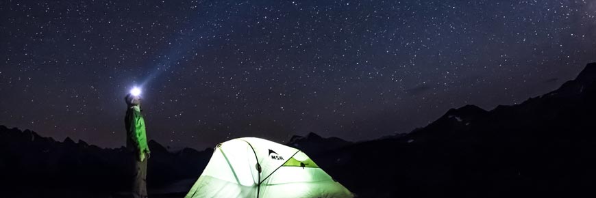 Un homme avec une lampe frontale regarde les étoiles devant sa tente.