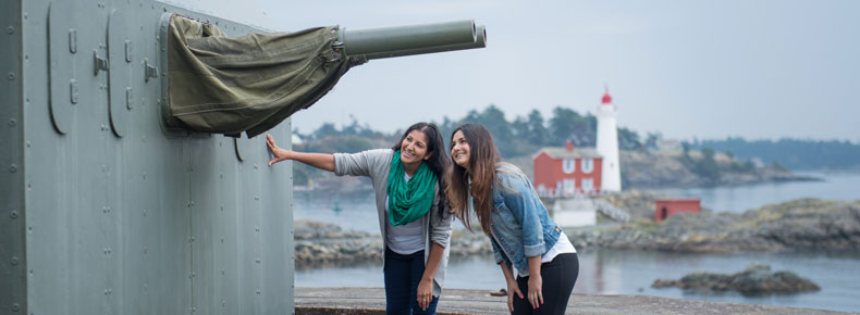 Deux jeunes femmes observent les canons bitubes de 6 livres à tir rapide de la Batterie Belmont avec le phare de Fisgard dans le fond.