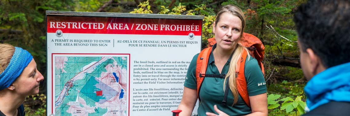 Un guide de Parcs Canada discute avec des randonneurs devant un panneau d’avis de fermeture avec la forêt en arrière-plan.