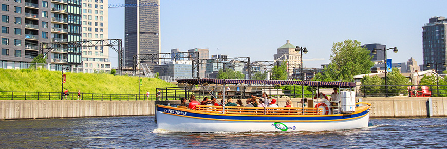 Un guide et des visiteurs sur le bateau Petit-Navire avec le canal de Lachine et la ville en arrière-plan