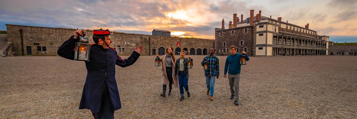 Quatre visiteurs et un guide avec des lanternes marchent dans la cour intérieure de la citadelle d’Halifax à la tombée de la nuit.