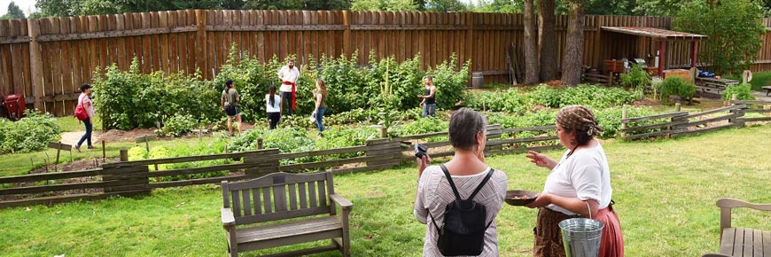 Un visiteur discute avec une jardinière en costume d’époque devant le jardin patrimonial du Fort-Langley pendant qu'un guide accompagne des enfants dans le jardin.