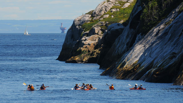 Des kayakistes dans le parc marin du Saguenay–Saint-Laurent avec des falaises, un bateau et un phare en arrière-plan.