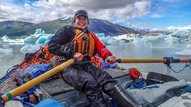 Un femme sourit lors d’une sortie de rafting sur la rivière Alsek à Kluane avec des montagnes en arrière-plan ainsi que des blocs de glace qui flottent.