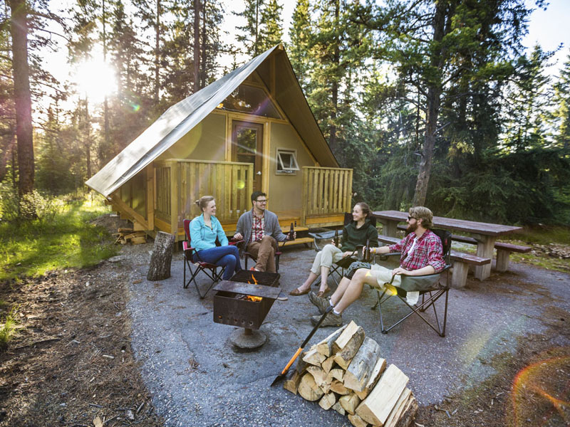 Un groupe de jeunes adultes autour d’un feu de camp, près d’une tente oTENTIK au terrain de camping Whistlers, au parc national Jasper.