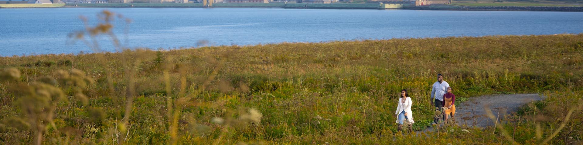 Trois adultes marchent dans un champ en bordure de la mer avec la forteresse de Louisbourg en arrière-plan.