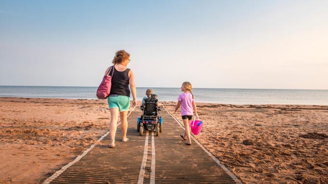 Une famille présentant des besoins en matière d’accessibilité marche en direction de l’eau sur la plage Stanhope en utilisant des tapis de mobilité.